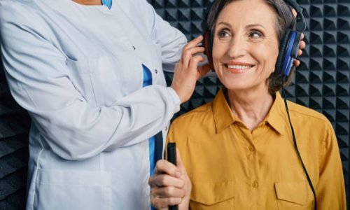ht2 Portrait senior woman with white toothy smile while hearing check-up with ENT-doctor at soundproof audiometric booth using audiometry headphones and audiometer